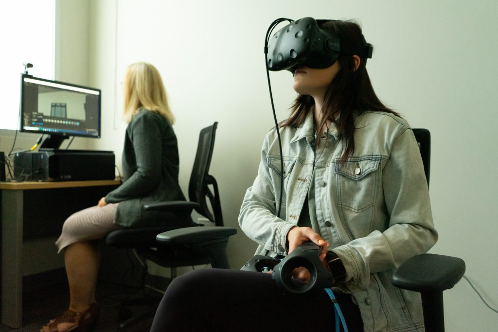 A student wearing a VR headset as a part of Deja Vu research looks around a virtual space. A researcher is seated in a chair monitoring what the student is viewing on a desktop computer.