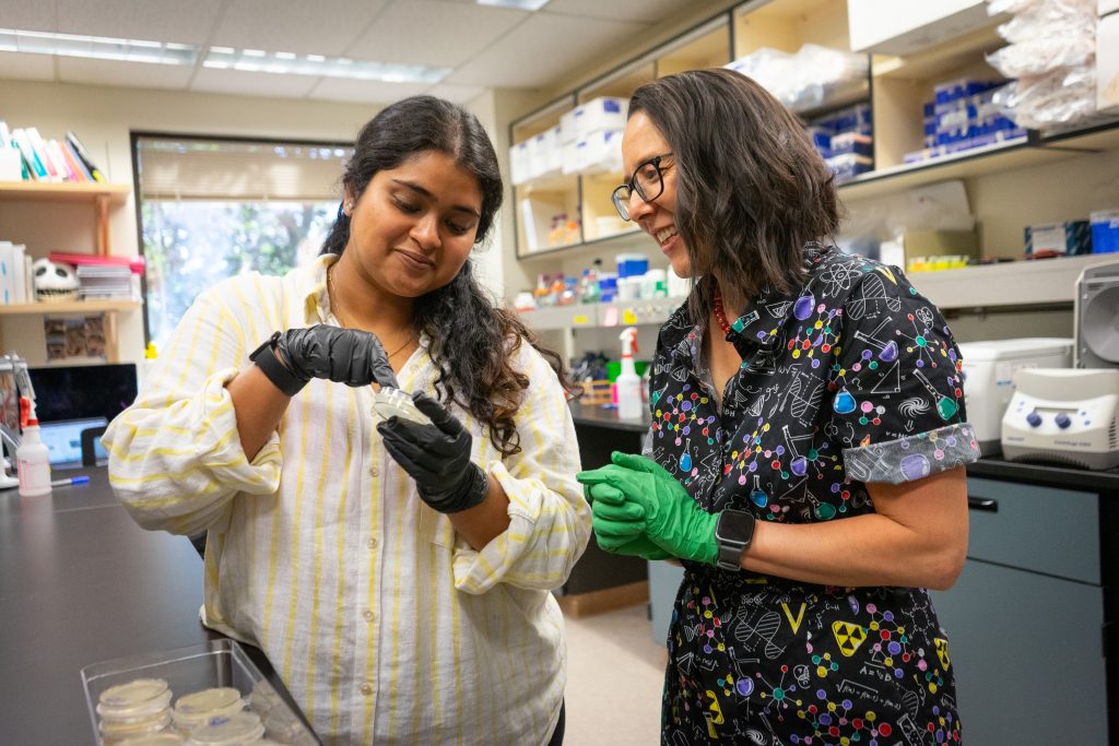 A graduate student in protective gloves holds a petri dish and points inside it as Erin Osborne Nishimura, a professor in a colorful science printed jumpsuit, looks on and smiles.