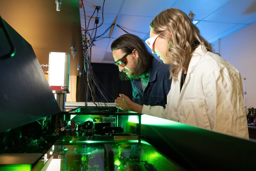 Two researchers in protective grasses look down at technical lab equipment which is tinged with green light.