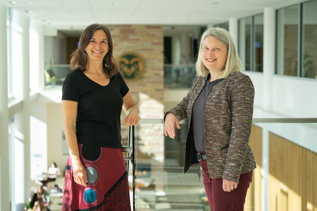 Two women smile and pose above the CSU Kindness Lounge, Kim Hoke on the left wearing a black t-shirt and maroon skirt, and Rachel Pries on the right wearing maroon pants and a blazer.