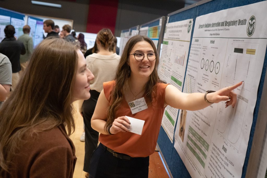 Statistics student Isabelle Munsell in an orange formal top presents her research at a poster session to a listening judge.