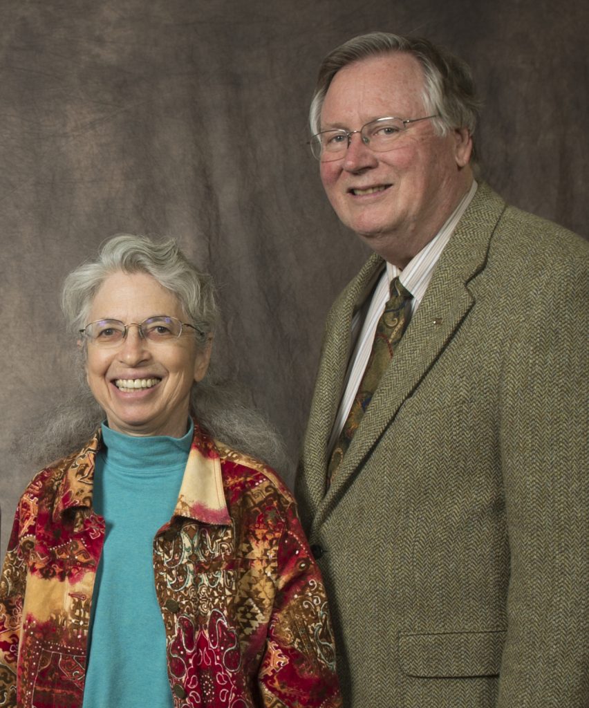 An older man and woman smile together in front of a grey studio backdrop. The woman wears a blue turtleneck and colorful red and yellow patterned coat. The man wears a brown suit and tie.