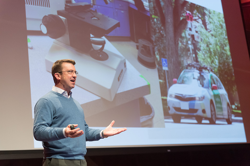 A man in a blue sweater and wearing a microphone talks in front of a projector screen displaying a Google Maps car. 